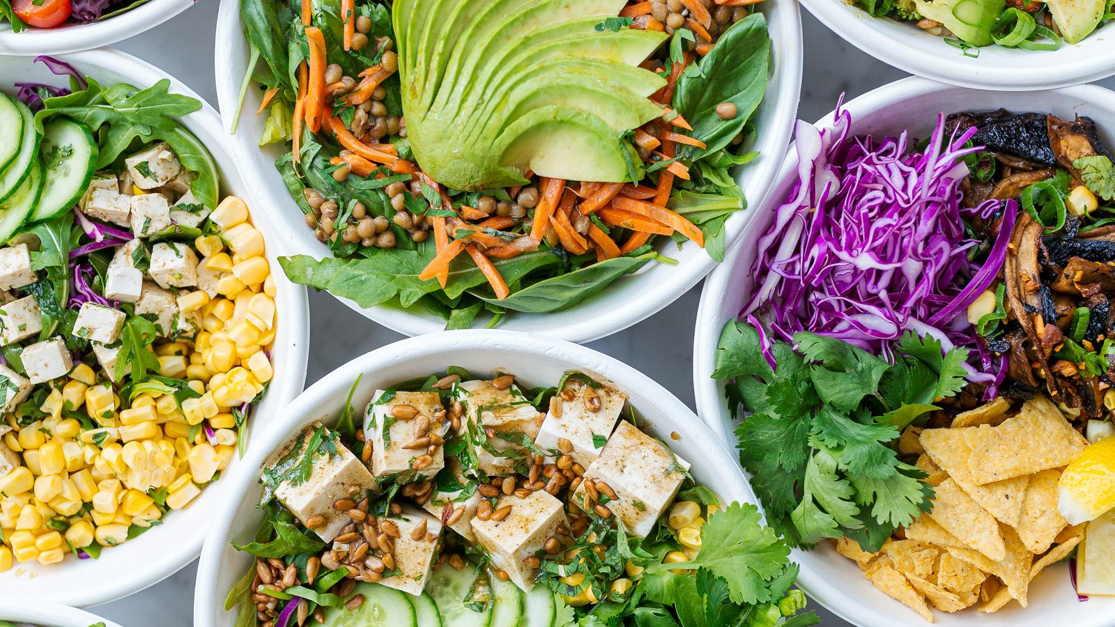 Overhead view of white bowls filled with salads: avocado and lentils, tofu and corn, cabbage and greens.